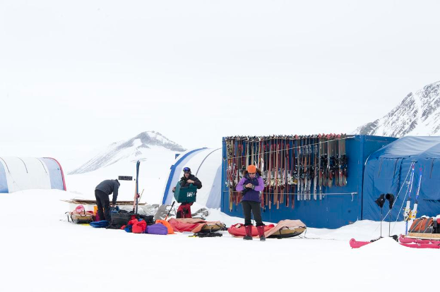 Drei Personen stehen auf einer schneebedeckten Landschaft mit Taschen drumherum, Zelte mit Skiern dahinter und schneebedeckte Hügel im Hintergrund unter einem klaren Himmel.