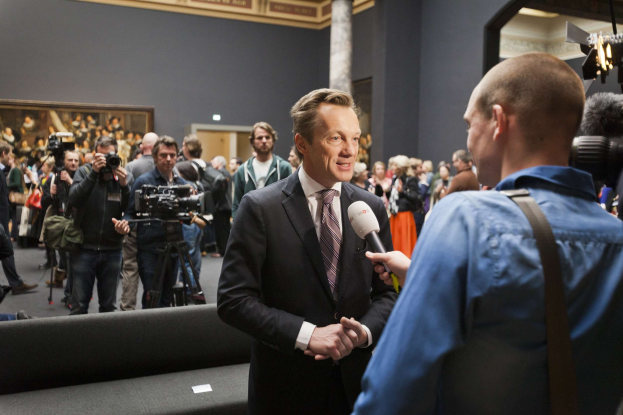 Ein Mann im Anzug hält ein Mikrofon und wird von einem Reporter vor einer Menge mit Kameras interviewt, ein Sofa im Vordergrund, ein gerahmtes Foto an der Wand und Beleuchtungsausrüstung.