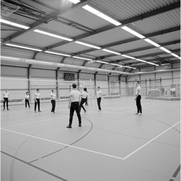 Eine Gruppe von Menschen, die Badminton in einer Turnhalle mit einem Netz in der Mitte und Lampen oben spielt, dargestellt in Schwarz-Weiß.