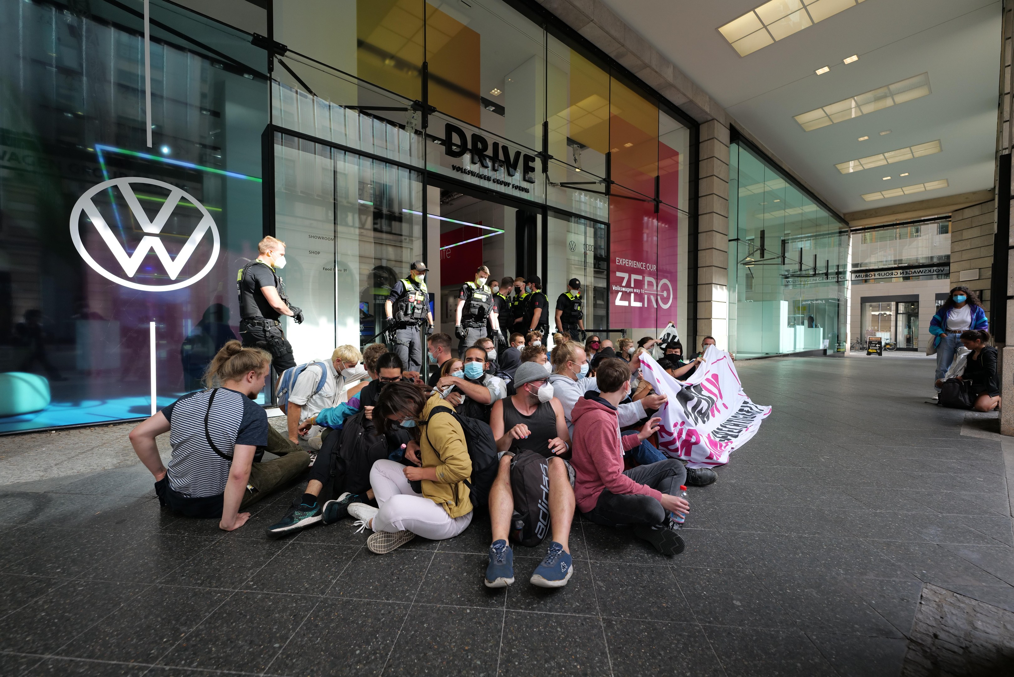 Eine Gruppe von Menschen sitzt vor einer Glasfassade eines Volkswagen-Händlers auf dem Boden und hält ein Transparent mit Text, unter Deckenleuchten, scheinbar protestierend.