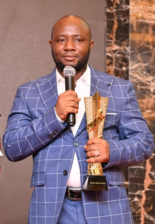 Ein Mann in einem blauen Anzug, der ein Mikrofon und eine Trophäe hält und selbstbewusst mit einem freudigen Gesichtsausdruck vor einer Wand steht.