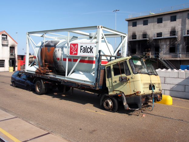 Ein Betonmischer-Lkw steht am Straßenrand, umgeben von Gebäuden mit Fenstern, Geländern und Treppen, mit Laternen und einem klaren blauen Himmel im Hintergrund.