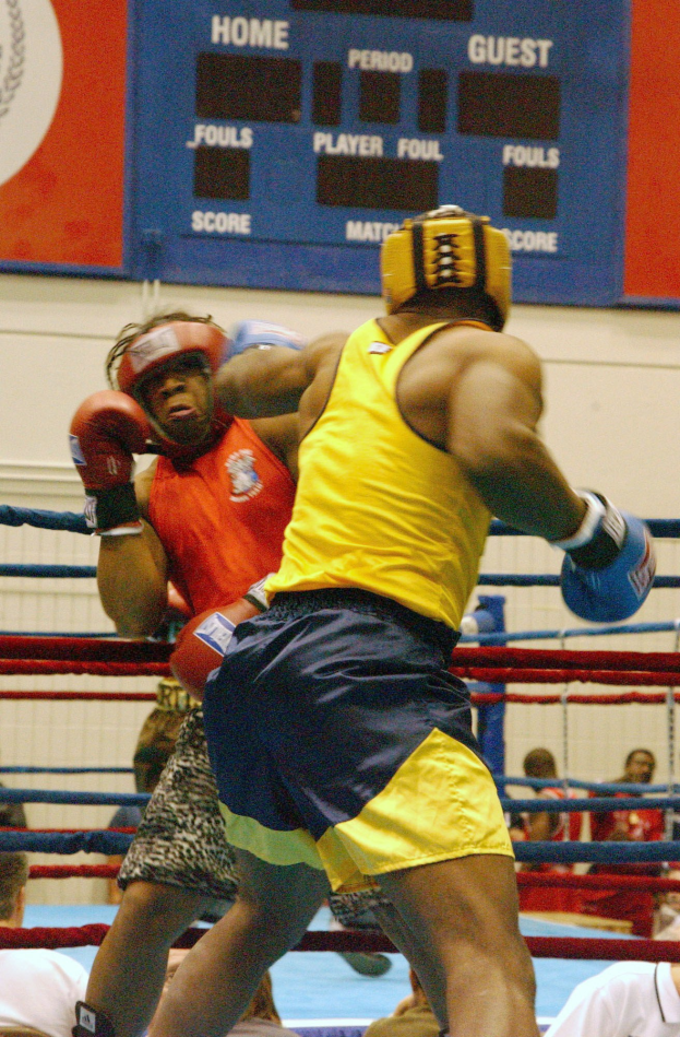 Zwei Boxer in einem Ring umgeben von einer Menge, mit einer Tafel im Hintergrund, die Text und Logos anzeigt.