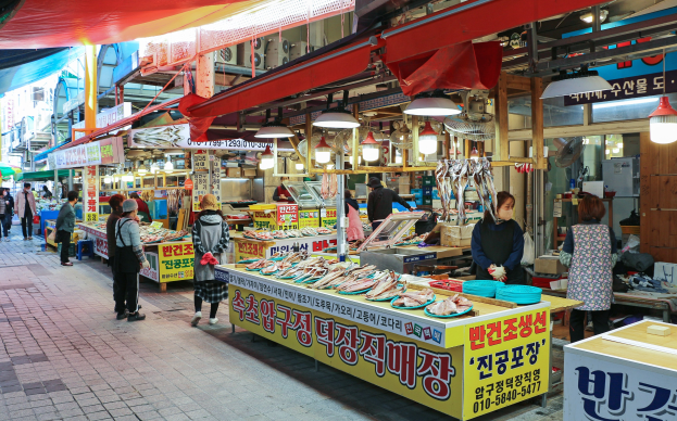 Ein belebter Straßenmarkt in Seoul, Südkorea mit Menschen, Ständen mit verschiedenen Artikeln und Gebäuden unter einem klaren blauen Himmel.