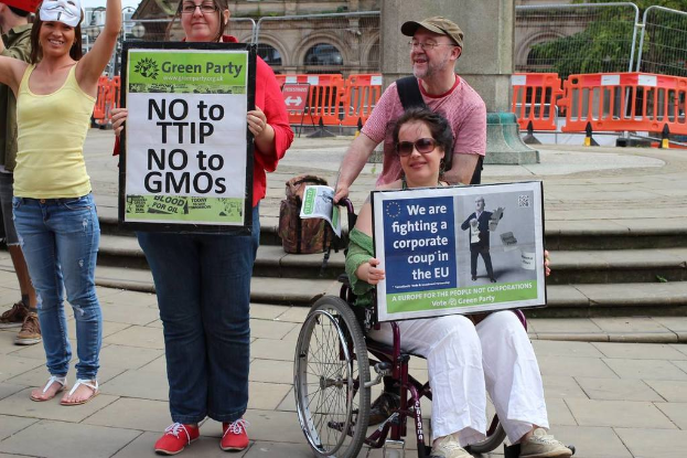 Eine Gruppe von Menschen in der Vordergrund, einige halten Plakate, mit einem Gehweg, einer Säule und einem Gebäude im Hintergrund.