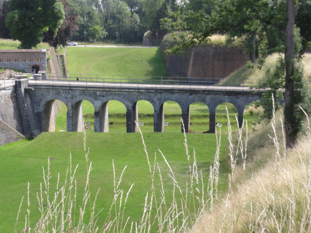 Eine steinerne Brücke mit Bögen und Säulen überspannt ein grünes Feld mit hohem Gras und Bäumen im Hintergrund, mit Fahrzeugen auf der Straße darunter und Geländern, die die Brücke umgeben.