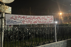 Ein Zaun mit einer Fahne, auf der 'Kashmirischer Protest gegen den Tod eines Mannes, der bei einem Autounfall starb' steht, vor einem Gebäude, umgeben von Motorrädern, Gras, Pflanzen, Straßenlaternen, Strommasten und Stromkabel unter einem klaren Himmel.