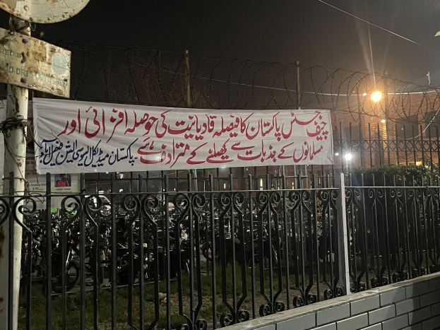 Ein Zaun mit einer Fahne, auf der 'Kashmirischer Protest gegen den Tod eines Mannes, der bei einem Autounfall starb' steht, vor einem Gebäude, umgeben von Motorrädern, Gras, Pflanzen, Straßenlaternen, Strommasten und Stromkabel unter einem klaren Himmel.