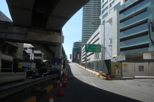 Eine Straßenansicht mit Absperrkegeln am Straßenrand, Fahrzeugen auf der Straße, einer Brücke auf der linken Seite, Gebäuden mit Schildern auf beiden Seiten, Laternen und einem Himmel im Hintergrund.