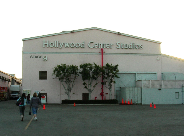 Hollywood Center Studios Gebäude mit Menschen vor dem Gebäude, Verkehrspfosten, Metallzaun, Bäume, Fahrzeuge, benachbarte Gebäude und bewölkter Himmel.