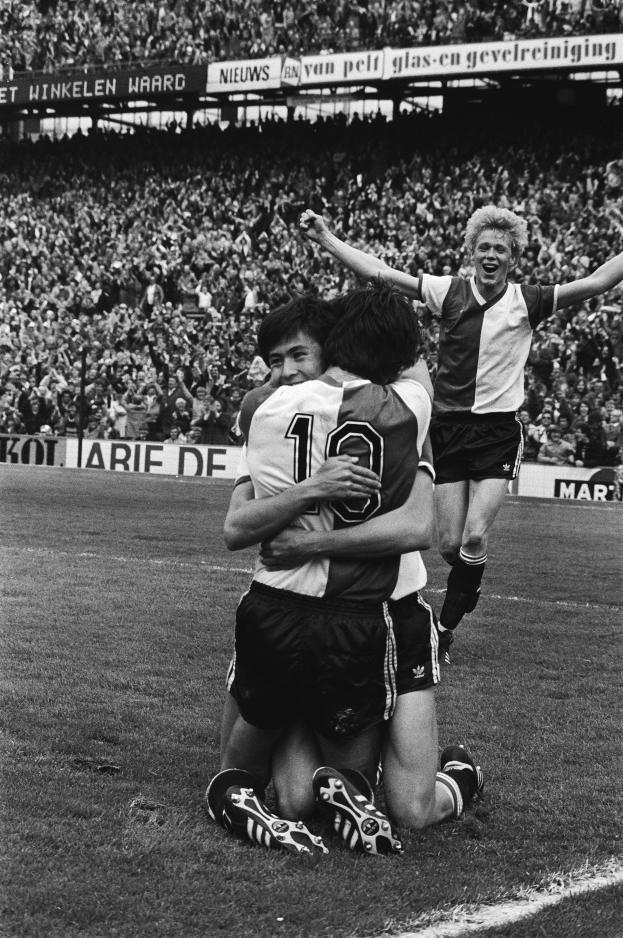 Drei Fußballspieler feiern ein Tor auf dem Boden, umgeben von jubelnden Fans und schwenkenden Fahnen im Hintergrund, dargestellt in Schwarz-Weiß.