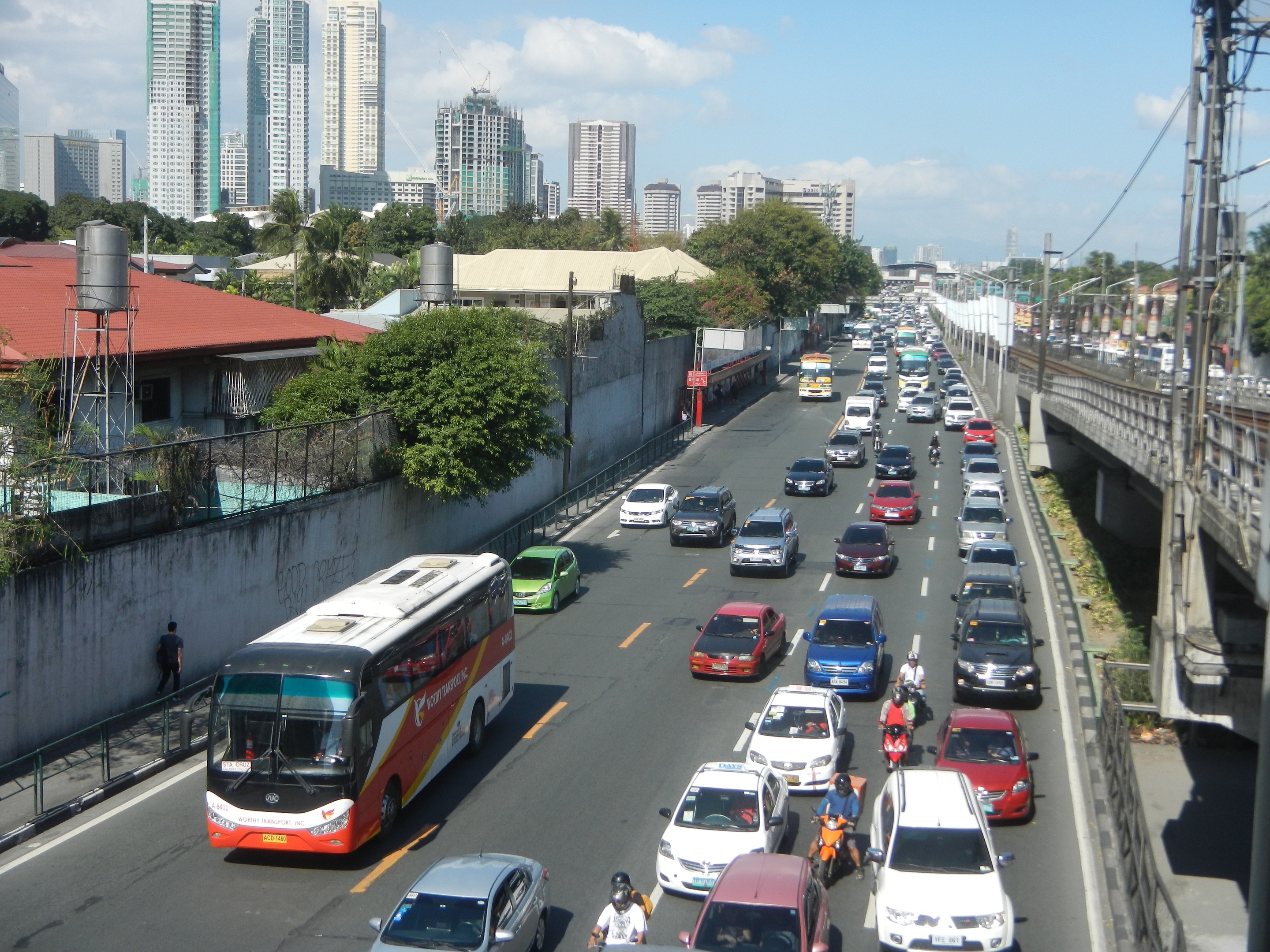 Eine belebte Stadtstraße mit Autos, Bussen und Motorrädern, die eine Brücke mit Strommasten und -leitungen, Gebäude, Bäume und einen bewölkten Himmel im Hintergrund zeigt.