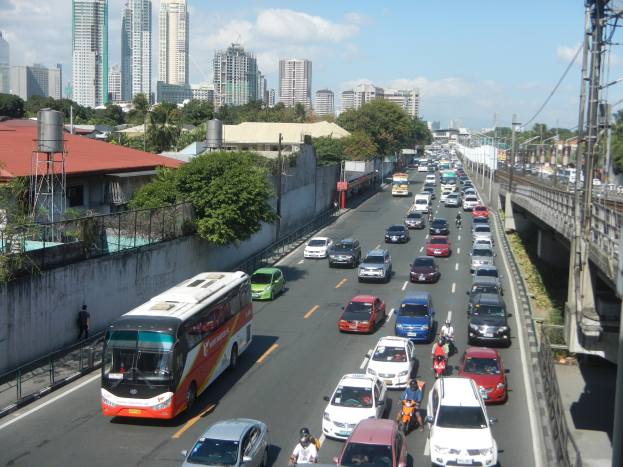 Eine belebte Stadtstraße mit Autos, Bussen und Motorrädern, die eine Brücke mit Strommasten und -leitungen, Gebäude, Bäume und einen bewölkten Himmel im Hintergrund zeigt.