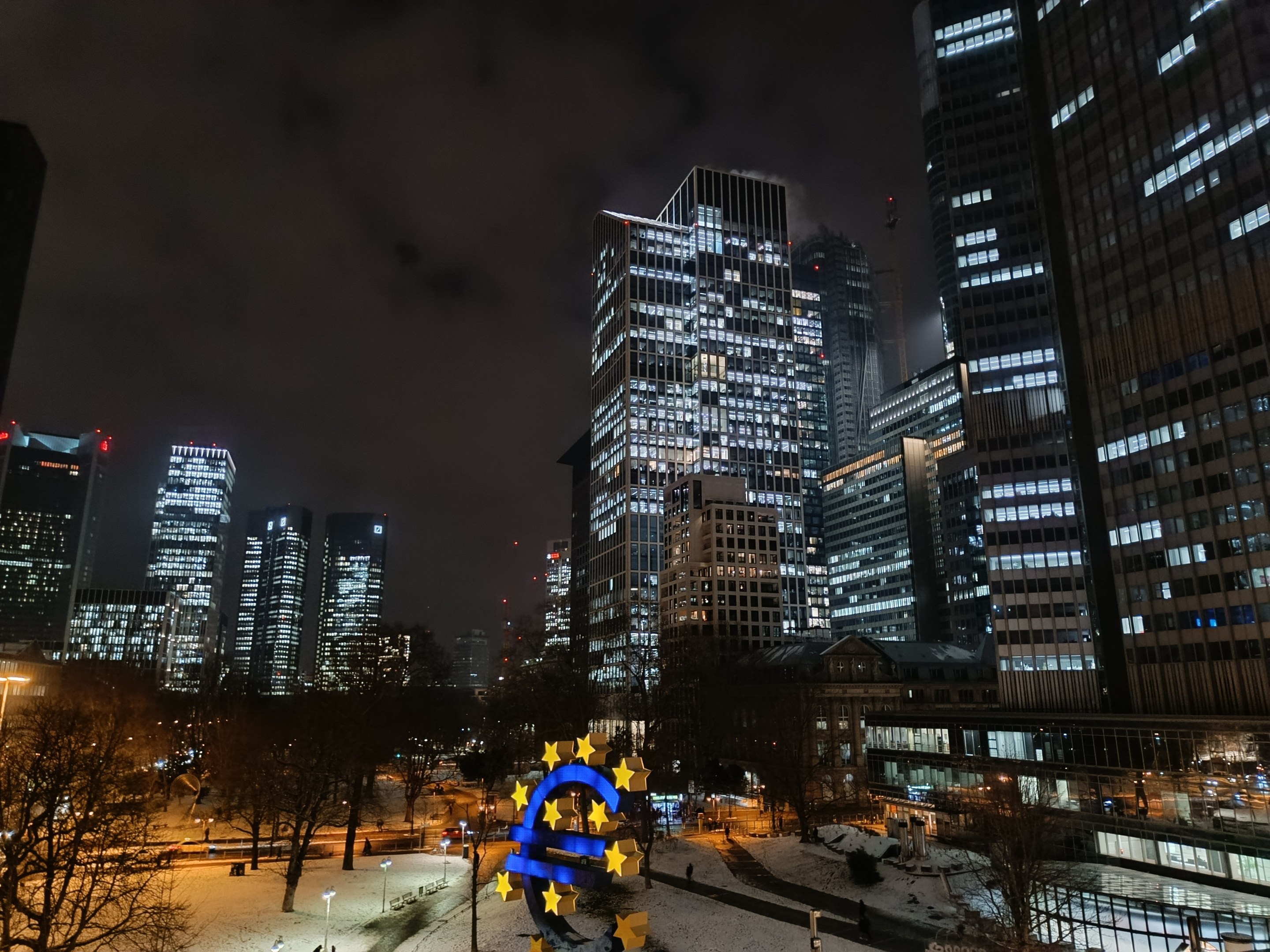 Europ├Ąische Zentralbank-Hauptsitz in Frankfurt, Deutschland bei Nacht, mit beleuchteten Geb├Ąuden, B├Ąumen, Stra├čenlaternen, Lichtern und Passanten unter einem bew├Âlktem Himmel.