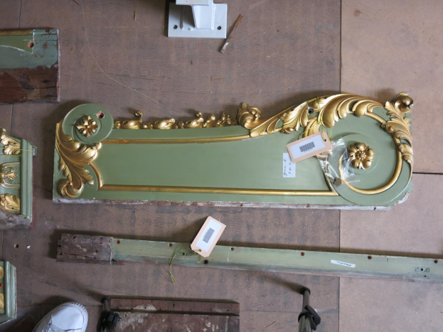 Ein vollgestellter Raum mit verschiedenen Möbelstücken, hölzernen Gegenständen, Etiketten und anderen Gegenständen, die auf dem Boden verstreut sind, mit einem Paar Schuhe unten, das auf eine Restaurierung hinweist.