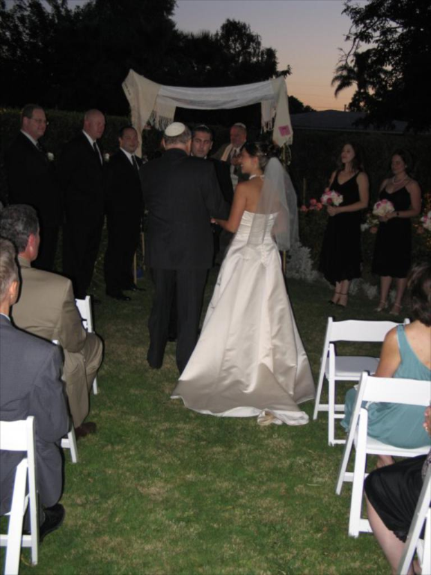 Eine Gruppe von Menschen steht bei einer Hochzeit, wobei die Frauen rechts Blumensträuße halten, während andere auf Stühlen sitzen und Bäume im Hintergrund zu sehen sind.