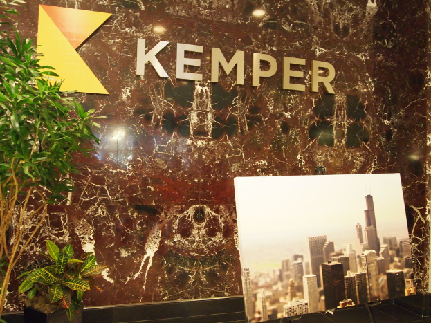 Eine Wand mit dem Wort "Kemper" darauf, flankiert von Pflanzen, mit einem Monitorbildschirm davor, der Gebäude zeigt.
