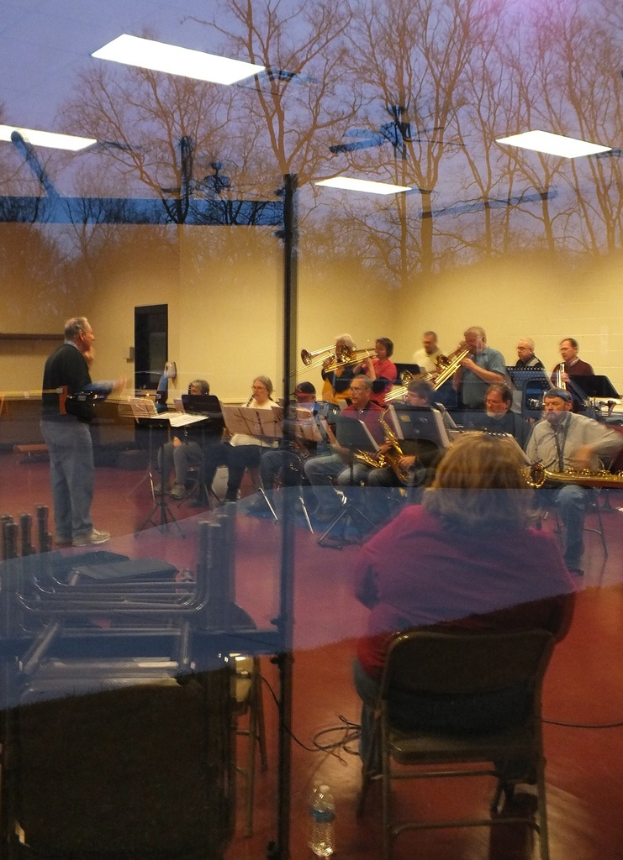 Musiker mit Trompeten im Hintergrund, Sänger vor Mikrofonen und eine Person, die Musik komponiert, mit einer Wand und Bäumen im Glasreflexion.