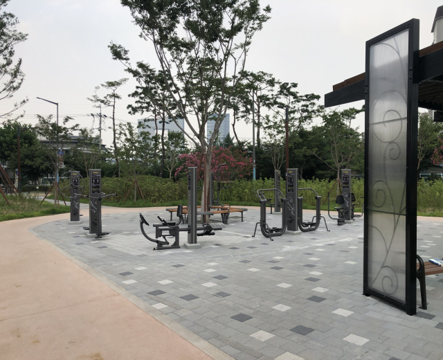 Outdoor park with fitness equipment, benches, trees, plants, grass, poles, lights, wires, and buildings under a sky backdrop.