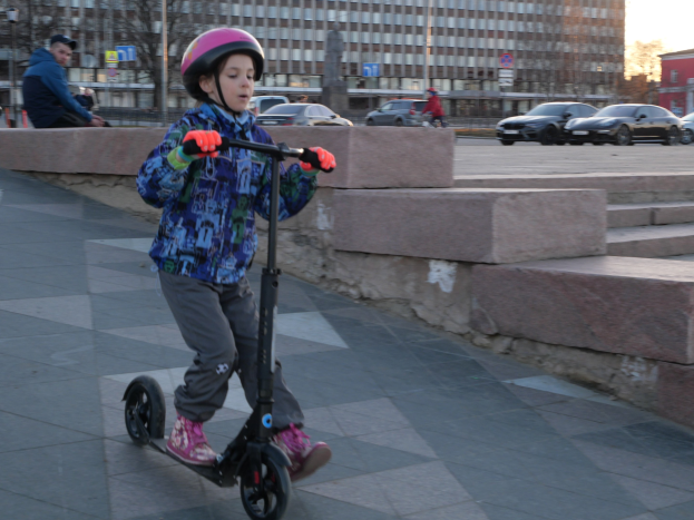 Ein junger Junge mit Helm und Handschuhen fährt auf einem Roller eine Treppe auf dem Gehweg hinunter, mit Fahrzeugen, Menschen, Bäumen, Pfählen, Brettern, Gebäuden und einem klaren blauen Himmel im Hintergrund.
