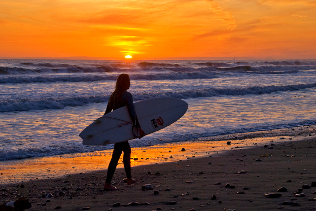 Eine Frau spaziert bei Sonnenuntergang am Strand entlang und hält ein Surfbrett in der Hand, mit einer felsigen Küste und einem farbenfrohen Himmel mit Wellen im Hintergrund.