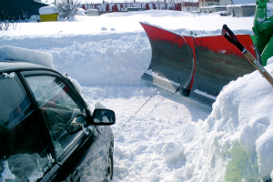 Ein Auto mit Schnee darauf, das auf einem schneebedeckten Boden mit verstreuten Gegenst├Ąnden geparkt ist, mit Geb├Ąuden und B├Ąumen im Hintergrund.