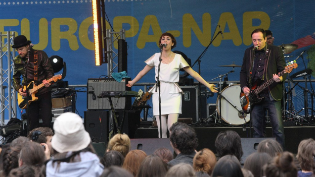 Vier Personen auf der Bühne: Eine Frau in einem weißen Kleid singt in der Mitte, während zwei Männer links und rechts Gitarren spielen; eine Menge ist unten sichtbar, mit Lautsprechern und einer Wand im Hintergrund.