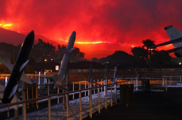 Ein Flugzeug fliegt über eine mit Raketen ausgestattete Landebahn, umgeben von Zäunen, Mülltonnen, Bäumen, Laternenmasten und Gebäuden, mit Rauch von den Kalifornischen Waldbränden am Himmel im Hintergrund.