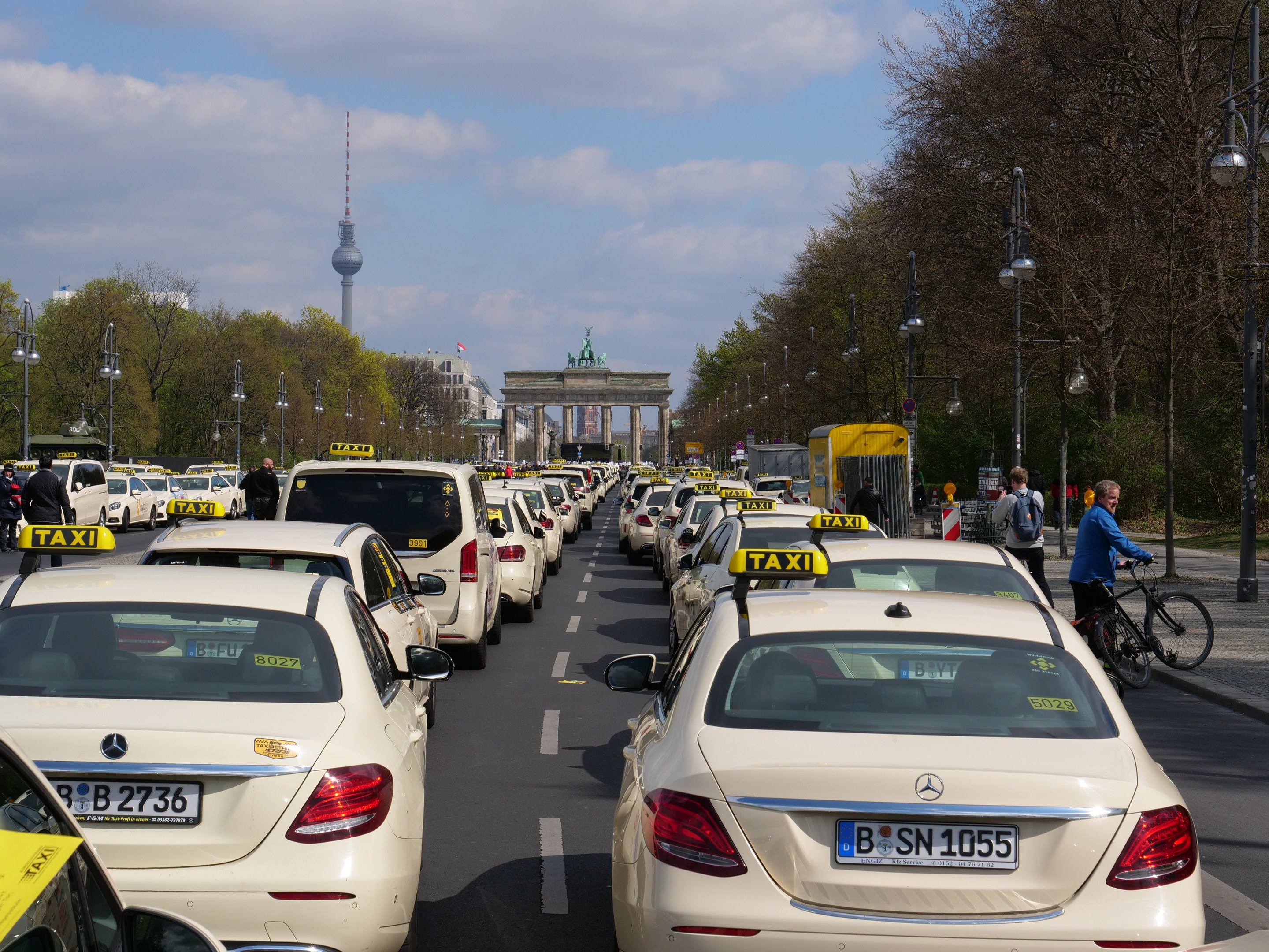 Eine lange Reihe von Taxis, die an einer belebten Straße in Berlin, Deutschland, geparkt sind, mit Fahrzeugen, Radfahrern und Fußgängern in der Nähe, flankiert von Laternenmasten, Bäumen und Gebäuden, einschließlich eines Bogens und eines Turms, unter einem bewölkten Himmel.