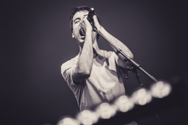 Ein Mann singt in ein Mikrofon auf einer hell erleuchteten Bühne, trägt ein schwarzes T-Shirt und Jeans und hat seine Haare wild gestylt.