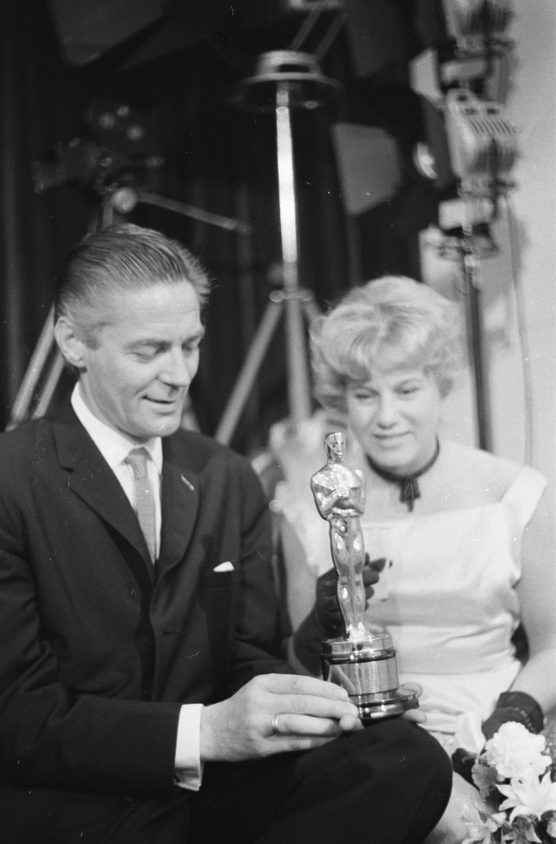 Ein Mann in einem schwarzen Blazer, weißem Hemd und Krawatte hält eine Oscar-Statue in der Hand und sitzt neben einer Frau, die einen Blumenstrauß hält, beide in festlicher Kleidung, mit Lichtern im Hintergrund, auf einem Schwarz-Weiß-Bild.