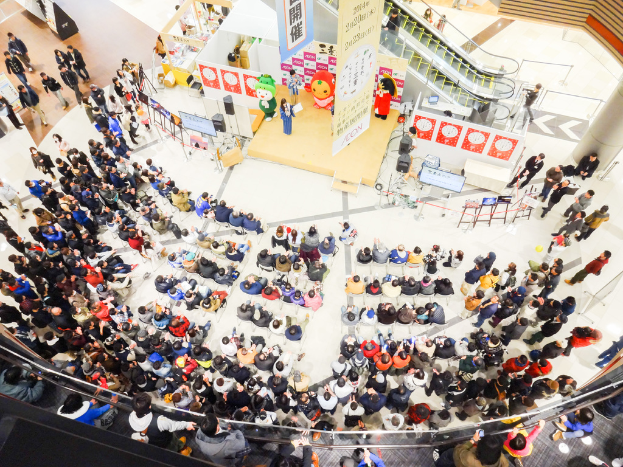 Eine Vogelperspektive eines Ereignisses mit vielen sitzenden und stehenden Menschen, einer Frau auf einem Podium, einer Rolltreppe dahinter und Tafeln neben dem Podium.