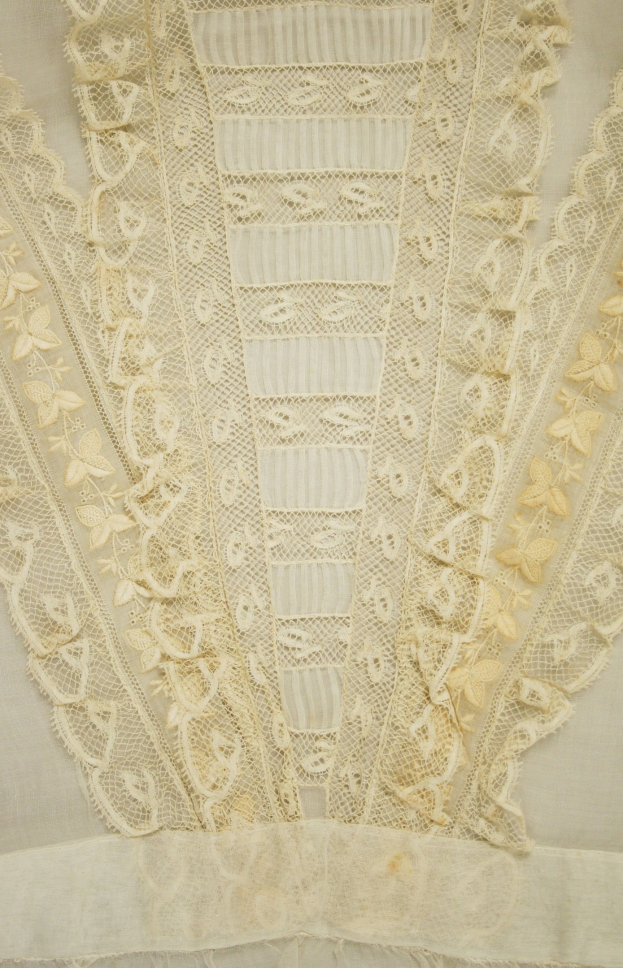 Nahaufnahme eines weißen Kleides mit zarter, kunstvoller Spitzeverzierung mit verschiedenen Formen und Mustern auf einem leichten, luftigen Stoff.