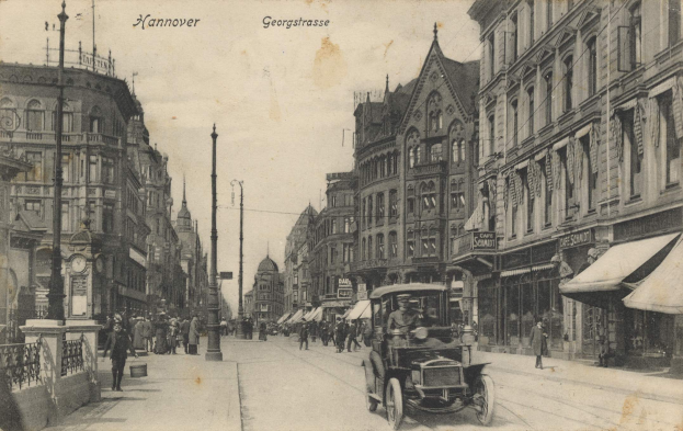 Ein altes Schwarz-Weiß-Foto einer Stadtstraße in Hannover, Deutschland, mit Fahrzeugen auf der Straße, Fußgängern auf den Gehwegen, Gebäuden mit Fenstern auf beiden Seiten, Pfählen, einer Uhr und einem sichtbaren Himmel.