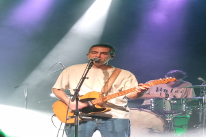 Ein Mann spielt Gitarre auf der Bühne bei einem Konzert, beleuchtet von Scheinwerfern über ihm, singt in ein Mikrofon, während ein anderer Mann hinter ihm Schlagzeug spielt.