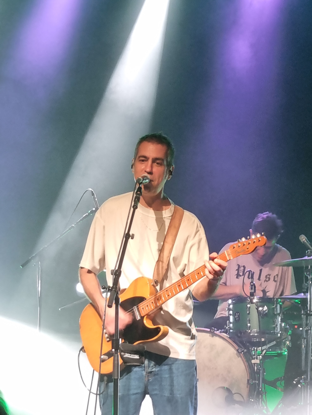 Ein Mann spielt Gitarre auf der Bühne bei einem Konzert, beleuchtet von Scheinwerfern über ihm, singt in ein Mikrofon, während ein anderer Mann hinter ihm Schlagzeug spielt.