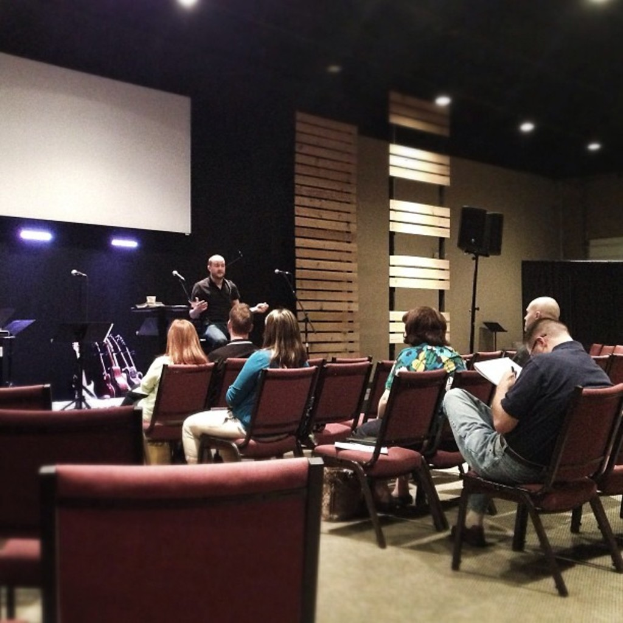 Innenansicht eines Saals mit Frauen auf roten Ledersesseln, einem Mann in schwarzem Hemd, der eine Rede hält, einem weißen Projektionsschirm, hölzernen Wanddekorationen, einem Lautsprecher auf einem Ständer und einer schwarzen Decke mit Spotlights.