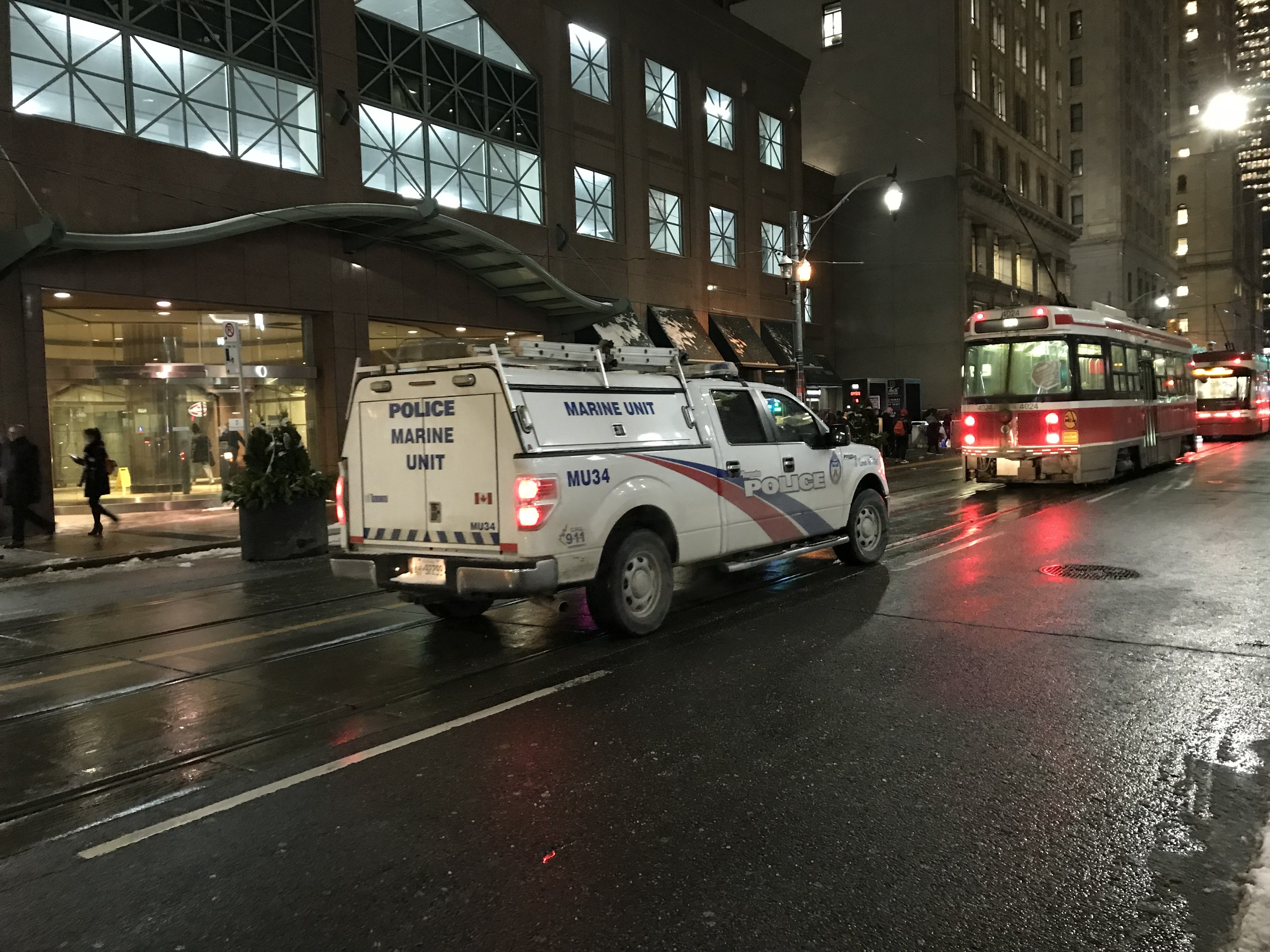 Ein Polizei-Lkw steht nachts am Straßenrand, mit Fahrzeugen auf der Straße, Fußgängern auf dem Gehweg, Topfpflanzen, Straßenlaternen, Gebäuden mit Fenstern und einem bewölkten Himmel.