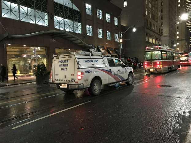 Ein Polizei-Lkw steht nachts am Straßenrand, mit Fahrzeugen auf der Straße, Fußgängern auf dem Gehweg, Topfpflanzen, Straßenlaternen, Gebäuden mit Fenstern und einem bewölkten Himmel.