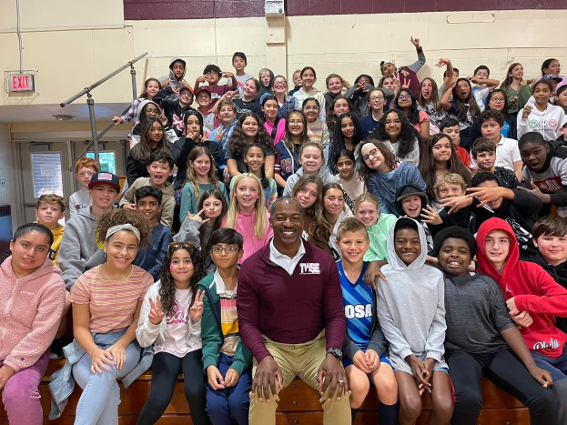Schüler posieren für ein Foto in einer Turnhalle bei einem Sommercamp, wobei einige auf der Treppe sitzen und andere stehen, eine Tür und ein Ausgangsschild links sichtbar