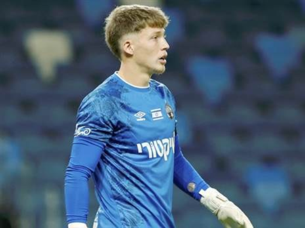 Ein junger Torwart in einem blauen Trikot steht auf einem Fussballfeld während eines Spiels, mit einem blauen T-Shirt und weißen Handschuhen.