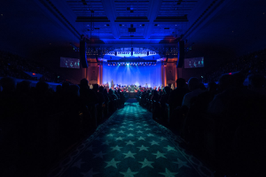 Ein großer Zuschauerraum mit Sitzenden, eine Bühne mit Musikern und eine Decke mit Lichtern und Fernsehgeräten, was auf einen Konzertsaal für die Nationale Convention 2019 hindeutet.