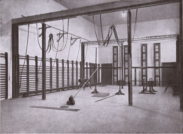 Schwarzes und weißes Foto eines Gymnastikraums mit verschiedenen Geräten wie Stangen und Seilen auf dem Boden, Fenstern im Hintergrund und Text unten.