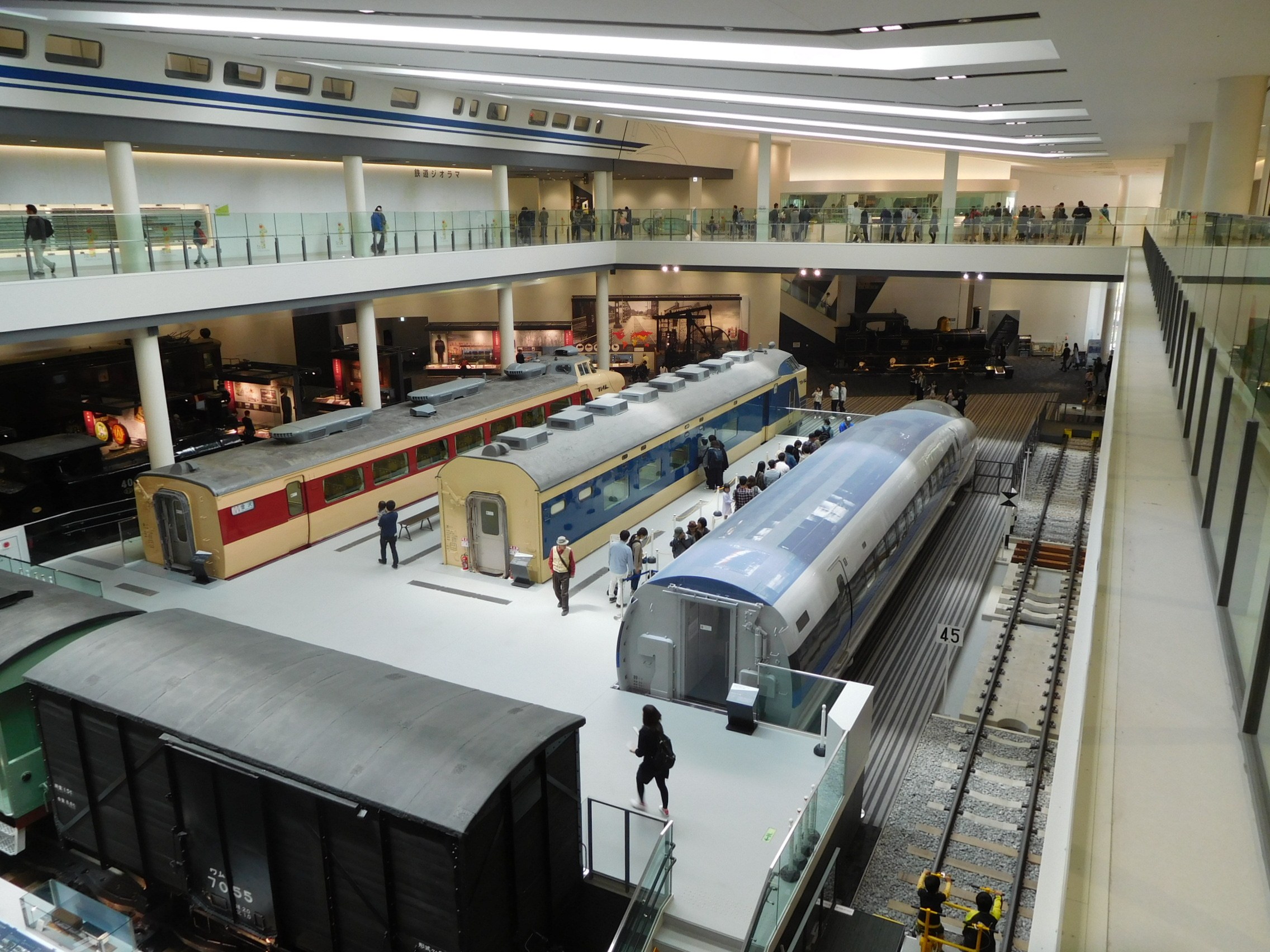 Großer Museumsraum mit verschiedenen Bahnen in unterschiedlichen Größen und Farben, mit Menschen, die beobachten, und Geländern, Säulen und Deckenbeleuchtung.