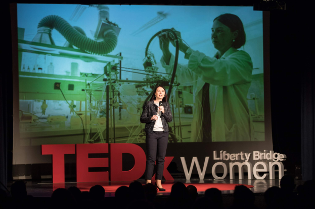Eine Frau steht auf einer Bühne und hält ein Mikrofon in der Hand, hinter ihr ist ein Bildschirm mit einem Bild einer Frau und Text, der sich auf ein TEDxWomen-Event bezieht.