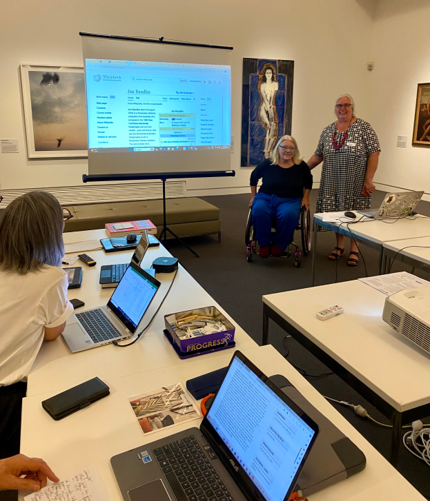 Eine Gruppe von Menschen, darunter eine Frau im Rollstuhl, sitzt um einen Tisch mit Laptops, Mobiltelefonen, Büchern und Papieren und nimmt an einem Workshop in einem Raum mit Fotorahmen und einer Projektionswand im Hintergrund teil.