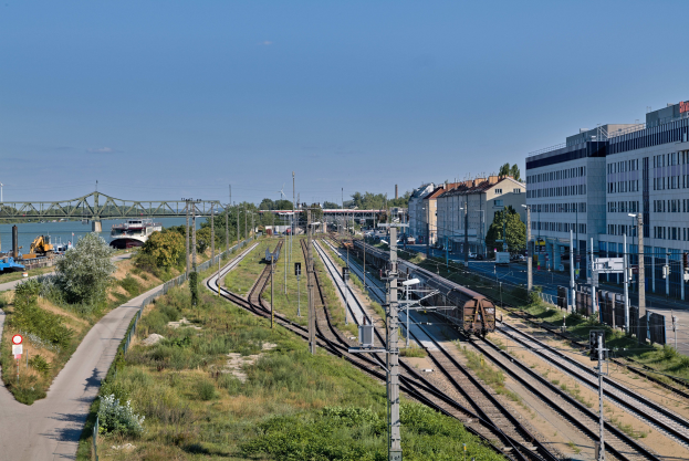 Ein Zug fährt auf Schienen neben einer Stadtlandschaft mit Gebäuden, Bäumen, Fahrzeugen, einer Brücke und elektrischen Oberleitungen unter einem bewölkten Himmel.