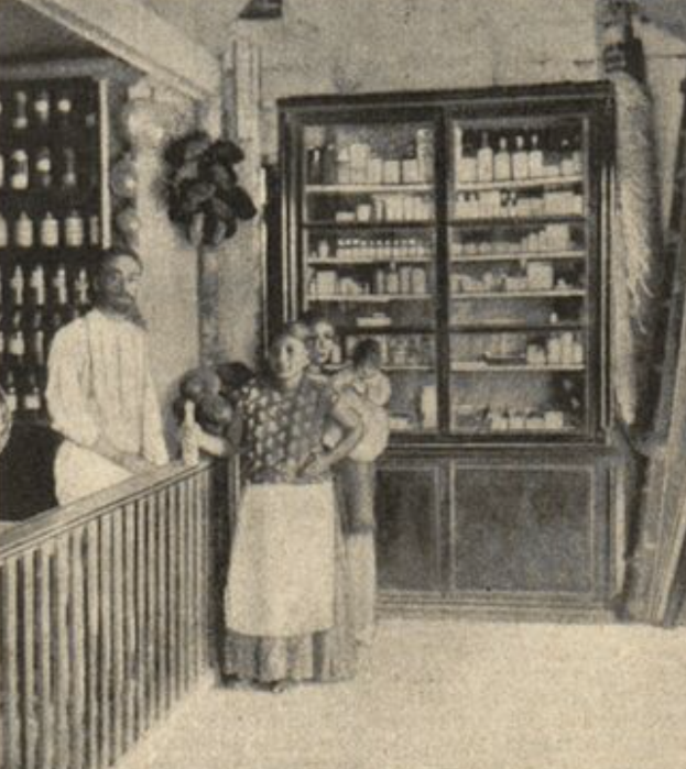 Ein altes Schwarz-Weiß-Foto einer Familie vor einer Apotheke mit einem Geländer auf der linken Seite und einem Schrank voller Flaschen im Hintergrund und Text unten.