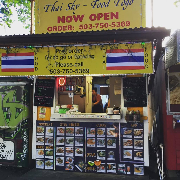 Ein Geschäft mit verschiedenen Artikeln wie Getränkedosen, Speisekarten, einer Uhr, Gewürzen in Plastikbehältern, Gewürzen, Werbetafeln und Namensschildern unter einem Himmel.