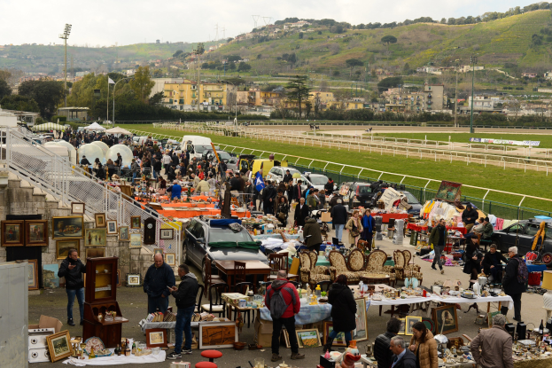 Eine große Gruppe von Menschen auf einem Flohmarkt mit Tischen, auf denen Gegenstände wie Rahmen und Stühle ausgelegt sind, Fahrzeuge in der Nähe geparkt, Geländer, Treppen, Bäume, Gebäude, Laternenpfähle, Hügel und ein bewölkter Himmel im Hintergrund.